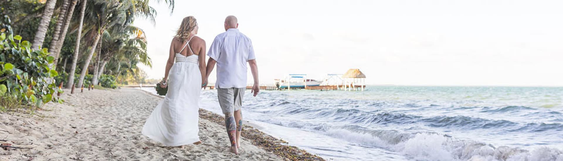 A blond bride in a gown and a bald groom with tattoos walk down the beach hand-in-hand.