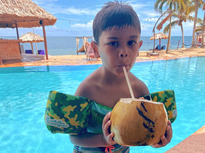 A young boy in swim floaties drinks from a coconut by a pool with a beach view.