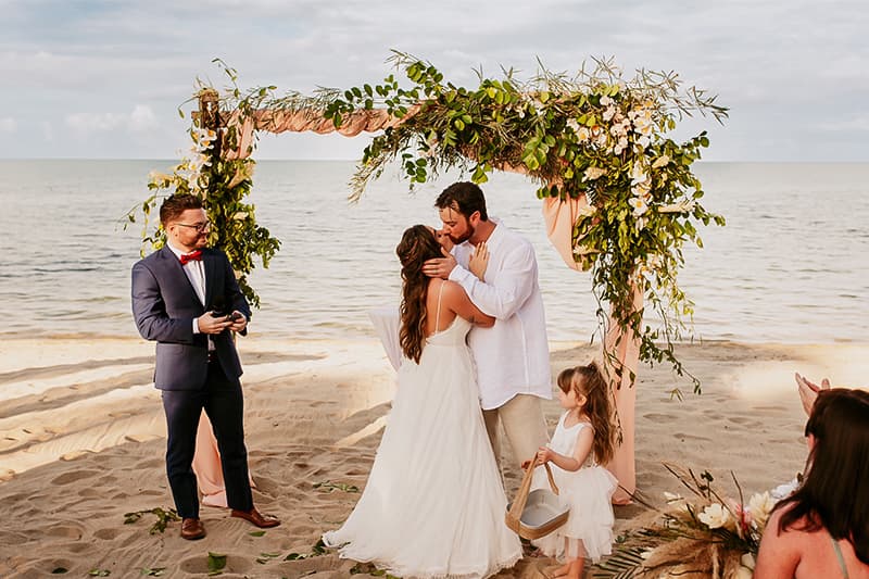 A bride and groom kiss under a floral arch on a beach, while a child holds a flower basket nearby. A bride and groom kiss under a floral arch on a beach, while a child holds a flower basket nearby.