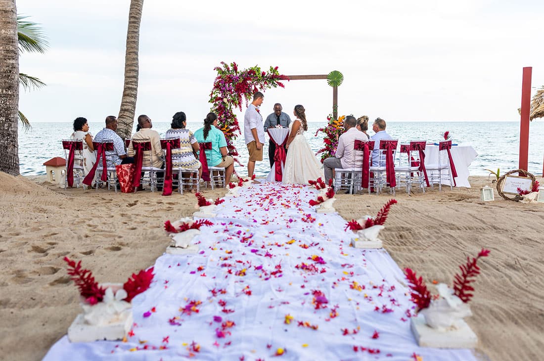 A couple are married on the beach under an arch decked out in flowers, surrounded by family and friends in clear acrylic chairs decorated with red satin bows.