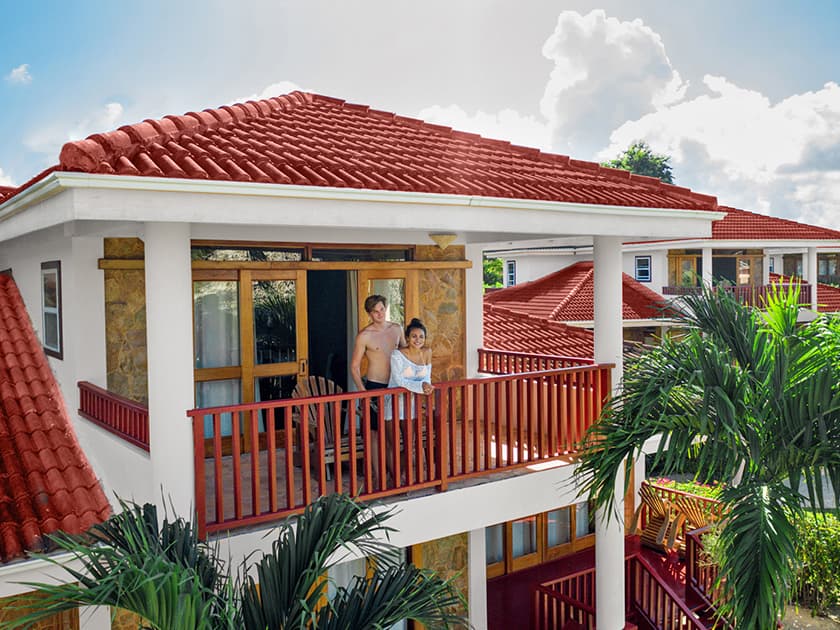 A couple stands on the balcony of a tropical villa with red roofs surrounded by palm trees.