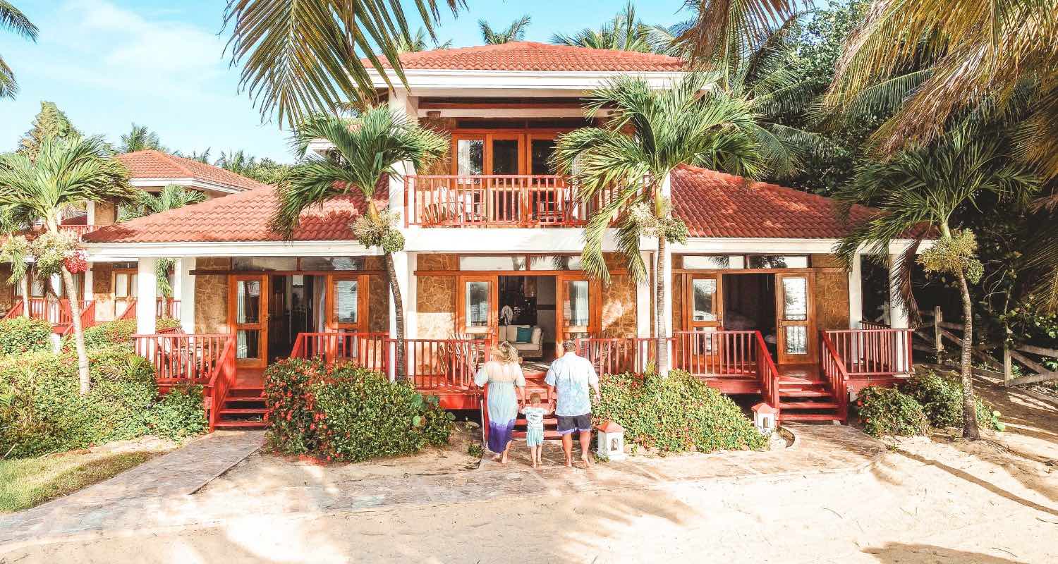 man and woman with 2 children walking up front stairs of their lodging building in the sand, with palm trees in the background.