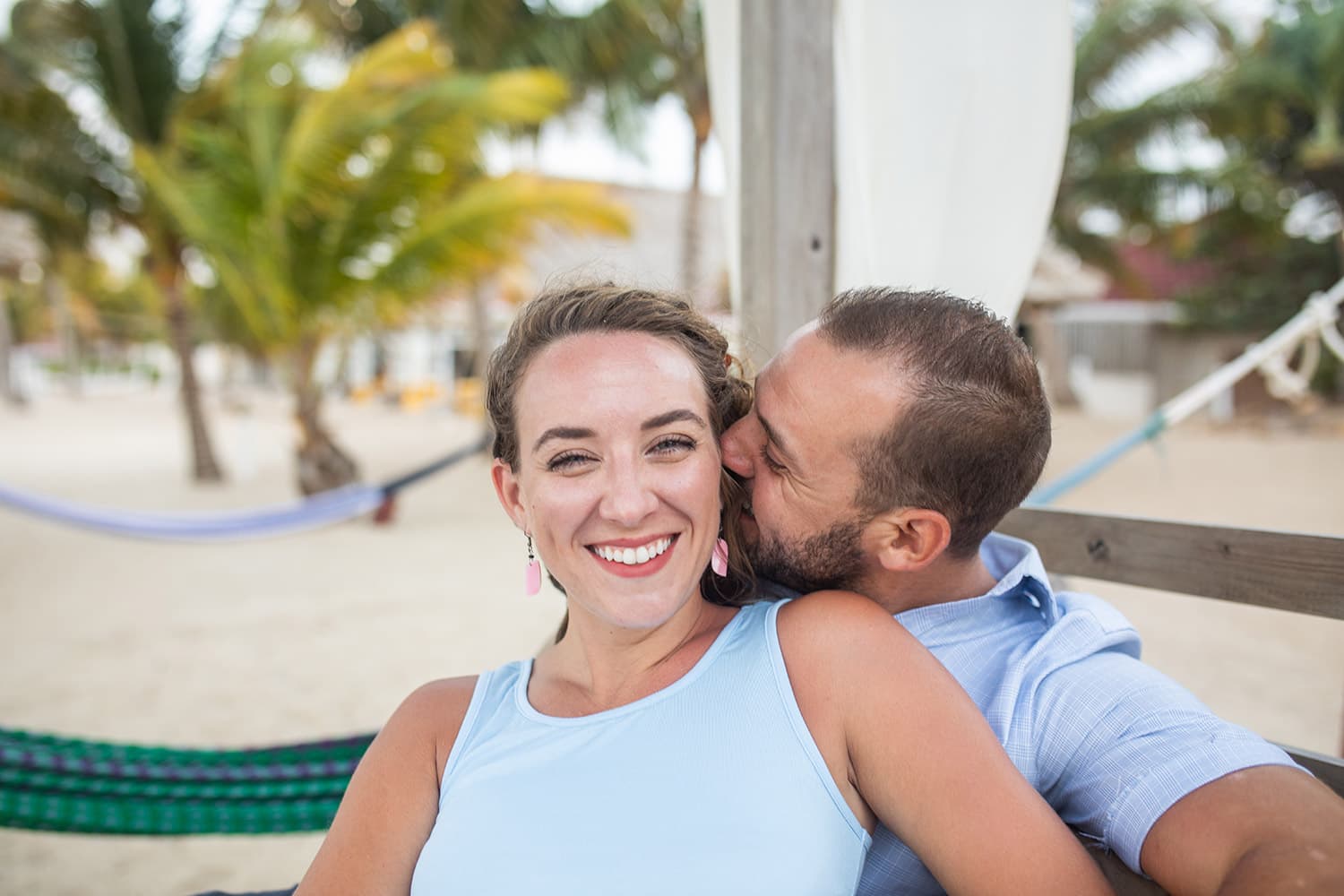 A dark-haired man playfully kisses a brunette woman on the ear surrounded by beach chairs and palm trees.