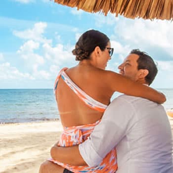 Brunet woman with glasses sitting on mans lap, under thatch umbrella on beach with ocean and sand in background.