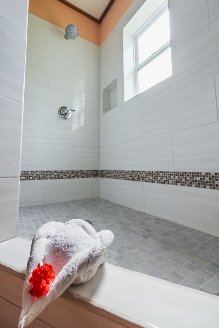 A modern, white tiled walk-in shower. The shower has a single showerhead, and there is a small window with a privacy glass panel. A mosaic tile border runs along the bottom of the shower, and the floor is tiled with small, square tiles.