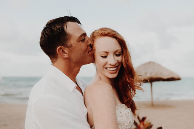 A couple shares an intimate moment on the beach, with the man kissing the woman's forehead as she smiles.