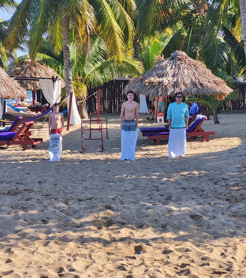 Three individuals standing on a sandy beach, each wrapped in white sacks, with palm trees and lounge chairs in the background.
