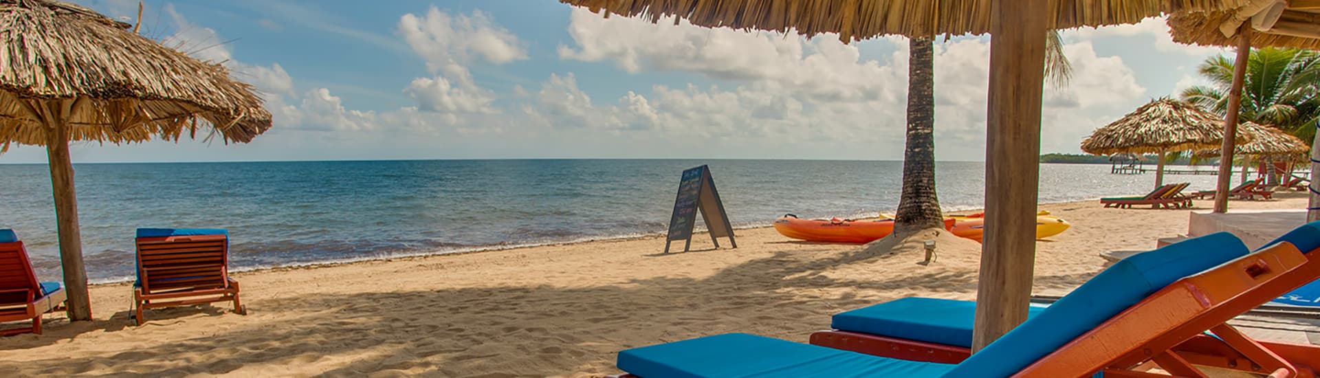 A sandy beach with wood lounge chairs with blue cushions under thatched roof umbrellas, overlooking the blue ocean, and a kayak and sandwich board sign on the far side of the beach.