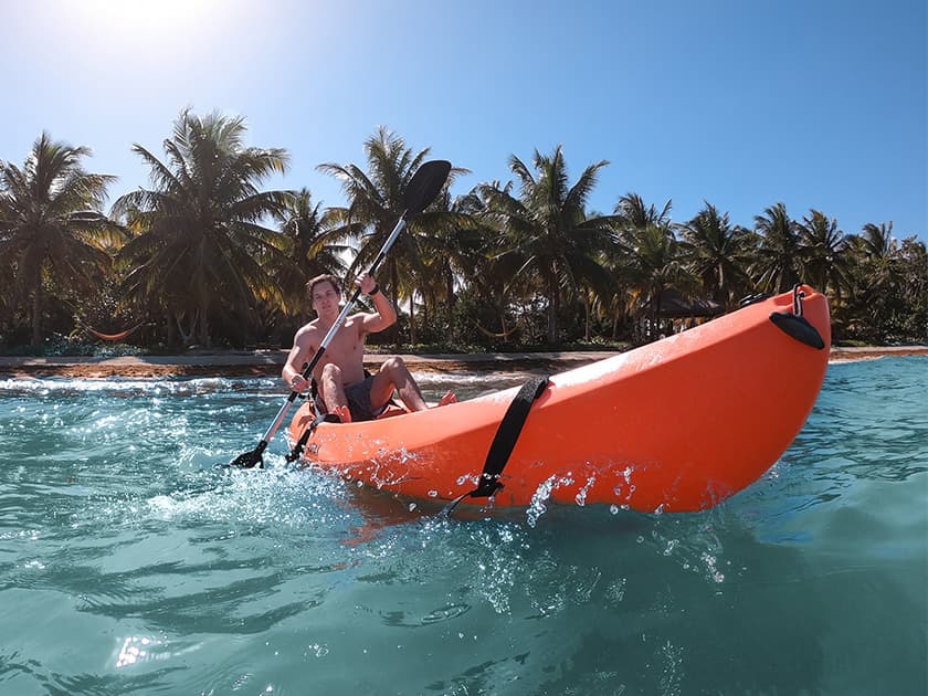 A person paddles an orange kayak in clear water surrounded by palm trees.