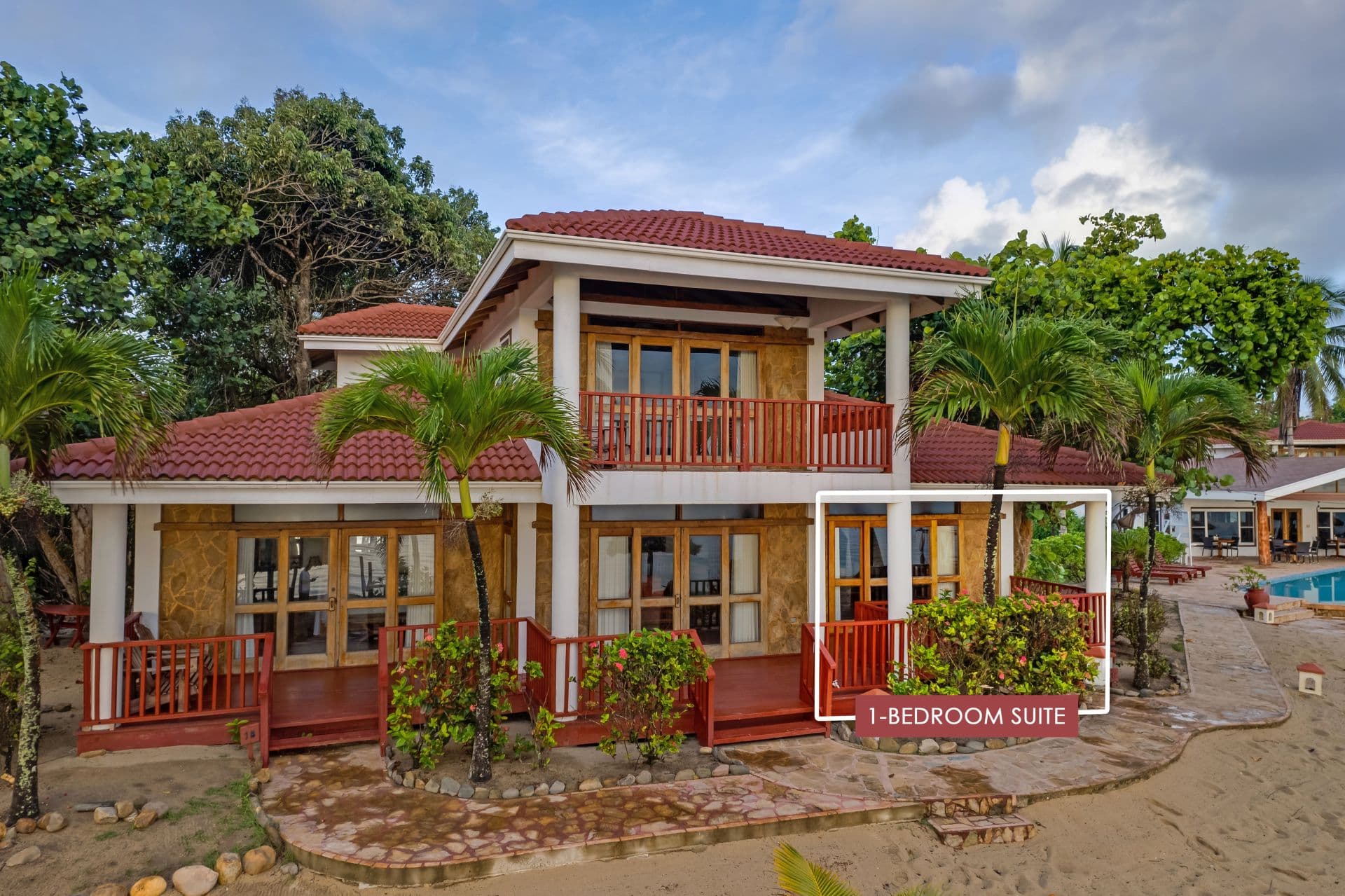 The image shows a 1-bedroom suite at Belizean Dreams Resort in Hopkins Bay, Belize. The suite is a two-story building with a red tile roof and a large balcony on the second floor. The suite is surrounded by palm trees and lush greenery.
