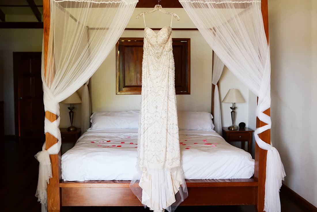 A white wedding dress hangs on a hanger in a four-poster bed with sheer white curtains. The bed is adorned with rose petals.