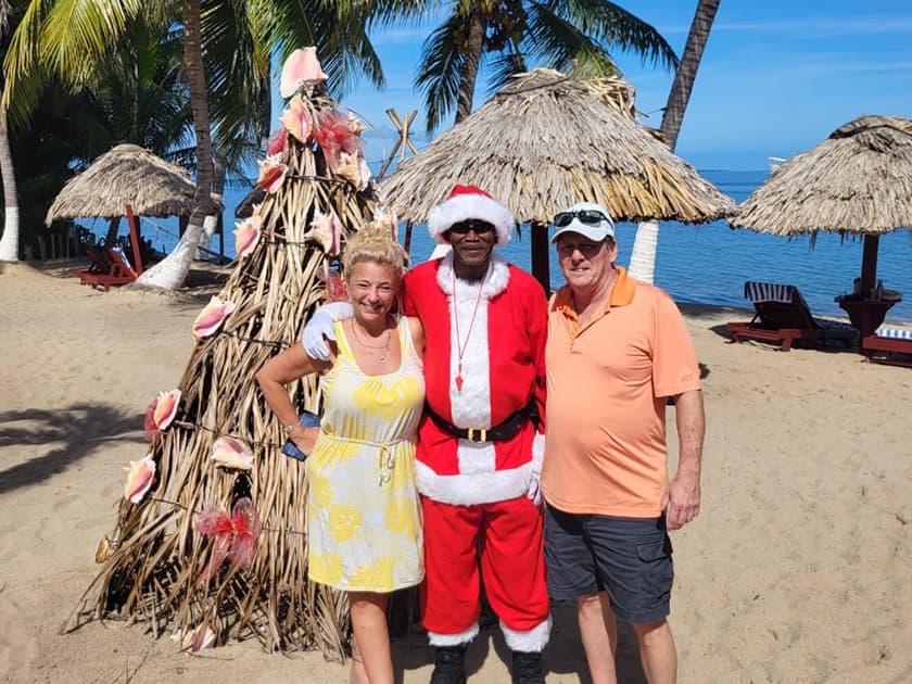 Three people, including a man in a Santa costume, pose together on a beach near a decorative palm tree structure. Three people, including a man in a Santa costume, pose together on a beach near a decorative palm tree structure.