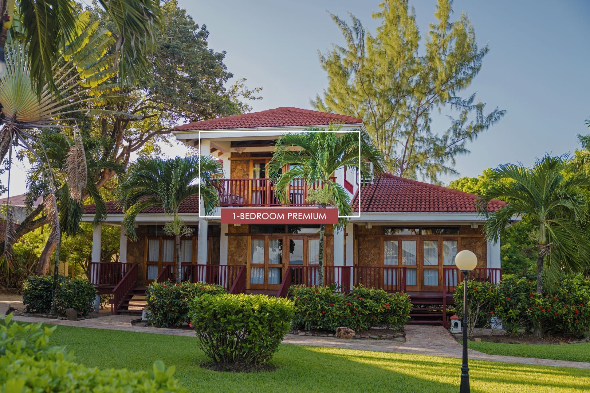The image shows a 1-bedroom premium room at Belizean Dreams Resort in Hopkins Bay, Belize. The room is a two-story building with a red tile roof and a large balcony on the second floor. The room is surrounded by palm trees and lush greenery.