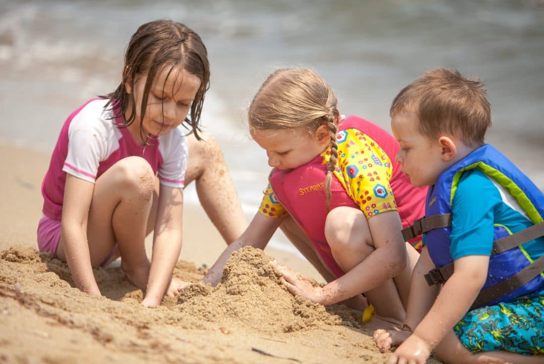 Three children in colorful bathing suits build castles in the sand on the beach.