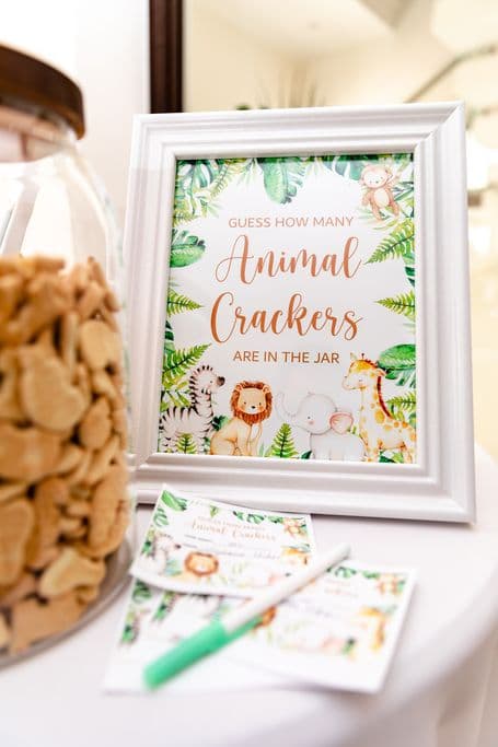 A white framed sign with the text "Guess How Many Animal Crackers Are In The Jar" and an illustration of various jungle animals. A clear jar filled with animal crackers sits next to the sign, and several blank cards and pens are placed on a table below.