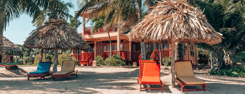 A beach scene featuring colorful lounge chairs and thatched umbrellas in front of a tropical house.