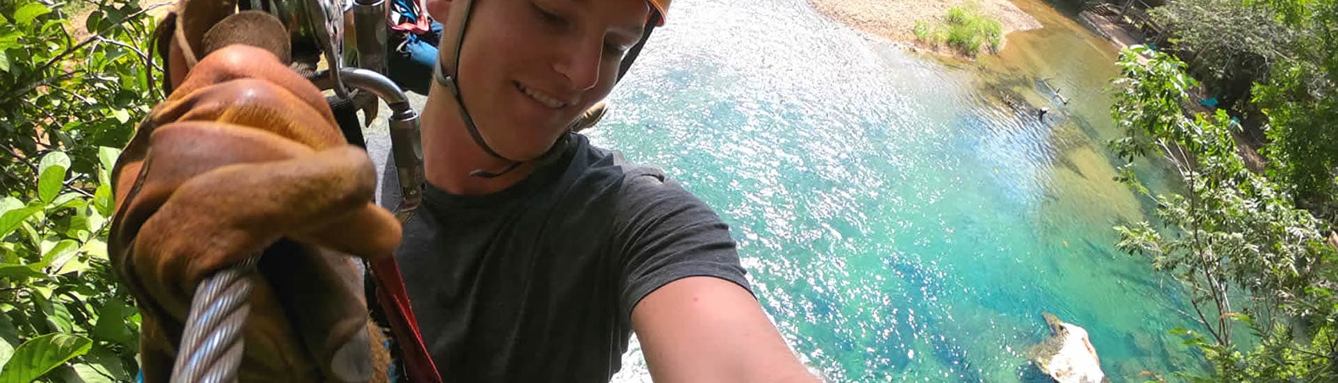 A smiling young man holds onto a zipline, with a leather glove, high above the water, zipping through the jungle.