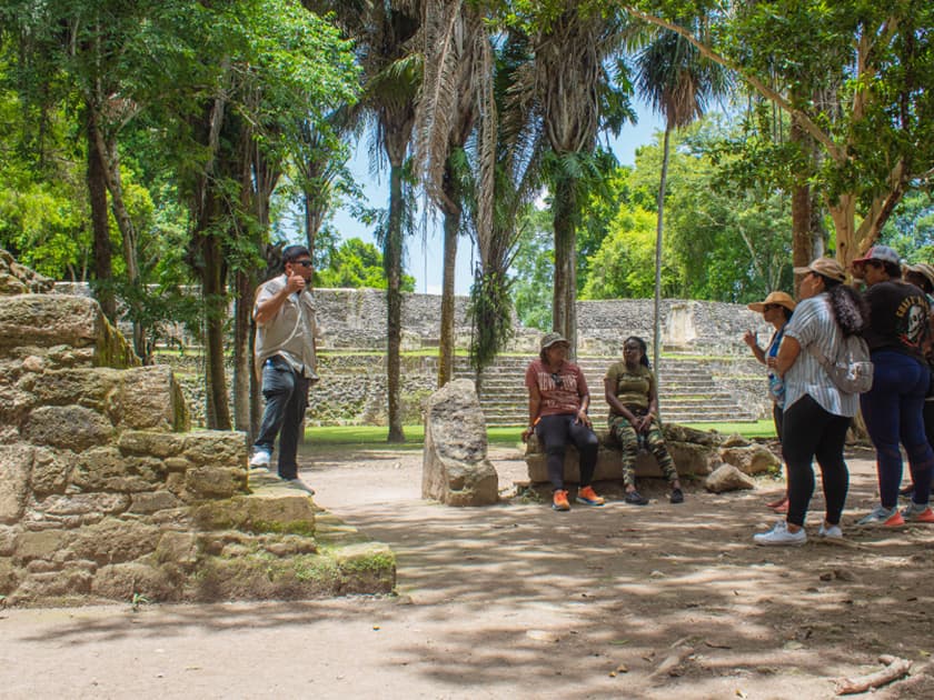 A tour guide speaks to a group of visitors near ancient ruins surrounded by lush greenery.