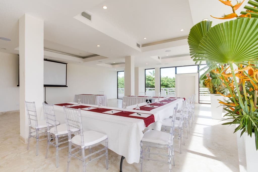 a spacious and modern meeting room with a U-shaped table arrangement. The room has a white tiled floor, white walls, and a high ceiling with exposed beams. There is a large palm tree in the corner, and a projection screen and projector at the front of the room. The table is covered in white tablecloths and has red runners. Clear plastic chairs are arranged around the table. There is a bar area with counter stools in the background.