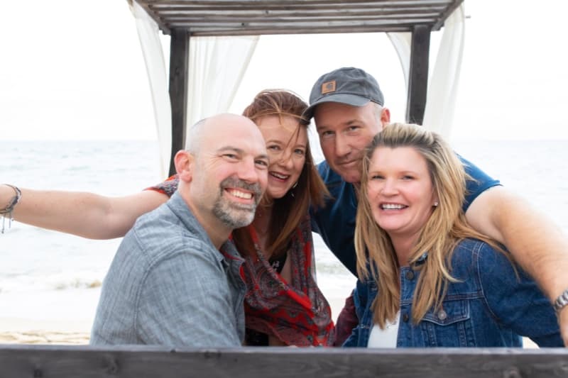 Four friends posing together under a wooden structure on the beach.