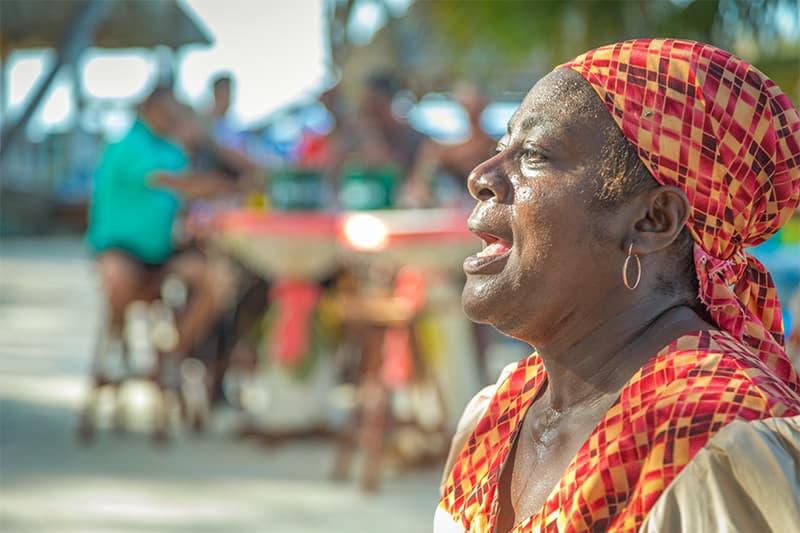 A woman in a colorful headscarf passionately sings outdoors, with a blurred background of people and vibrant scenery.