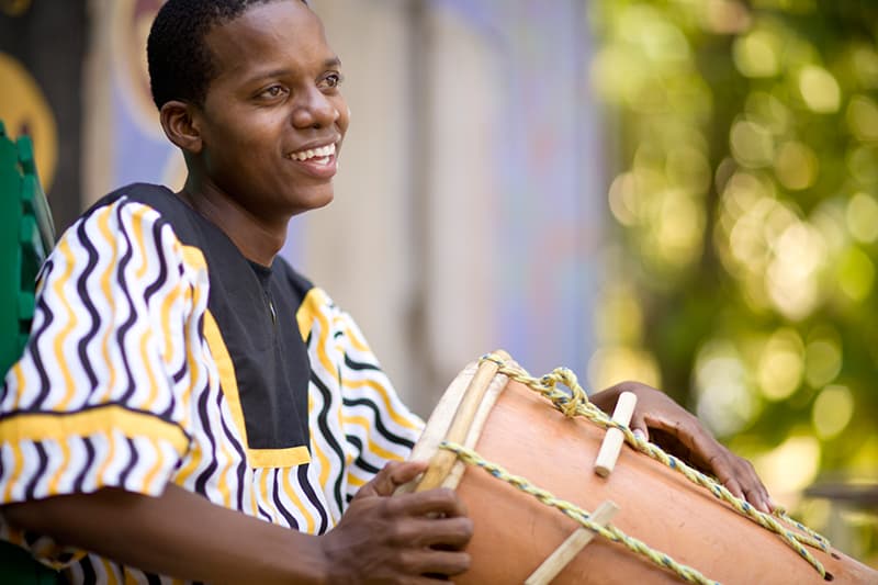 A smiling musician in traditional attire plays a hand drum outdoors. A smiling musician in traditional attire plays a hand drum outdoors.