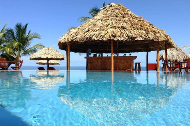A tranquil poolside view featuring a thatched-roof bar and sun loungers against a clear blue sky.