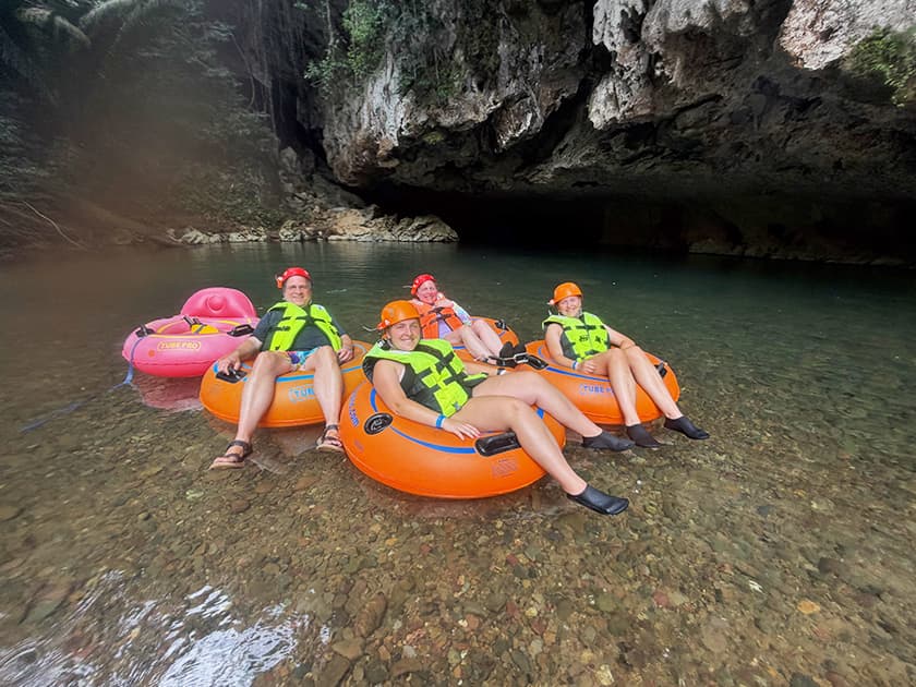 Five individuals in colorful inner tubes float on a calm river beneath a rocky overhang. Five individuals in colorful inner tubes float on a calm river beneath a rocky overhang.