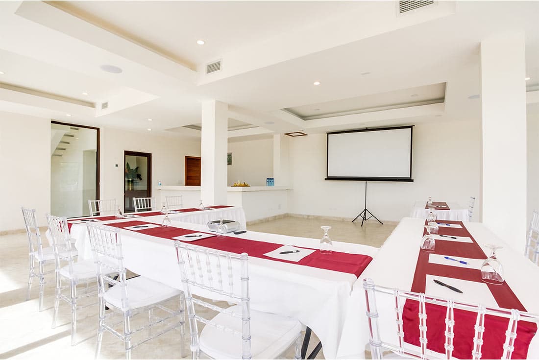 A modern meeting room with a U-shaped table arrangement, white tablecloths, and red table runners. There is a projection screen at the front of the room, and clear ghost chairs are provided for attendees.