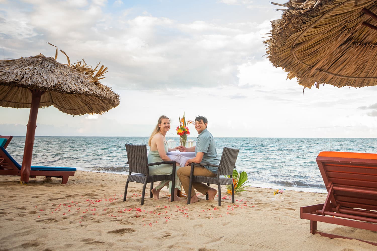 A woman in a green dress and a man in a blue shirt hold hands while sitting at a romantic dinner table placed on the beach.