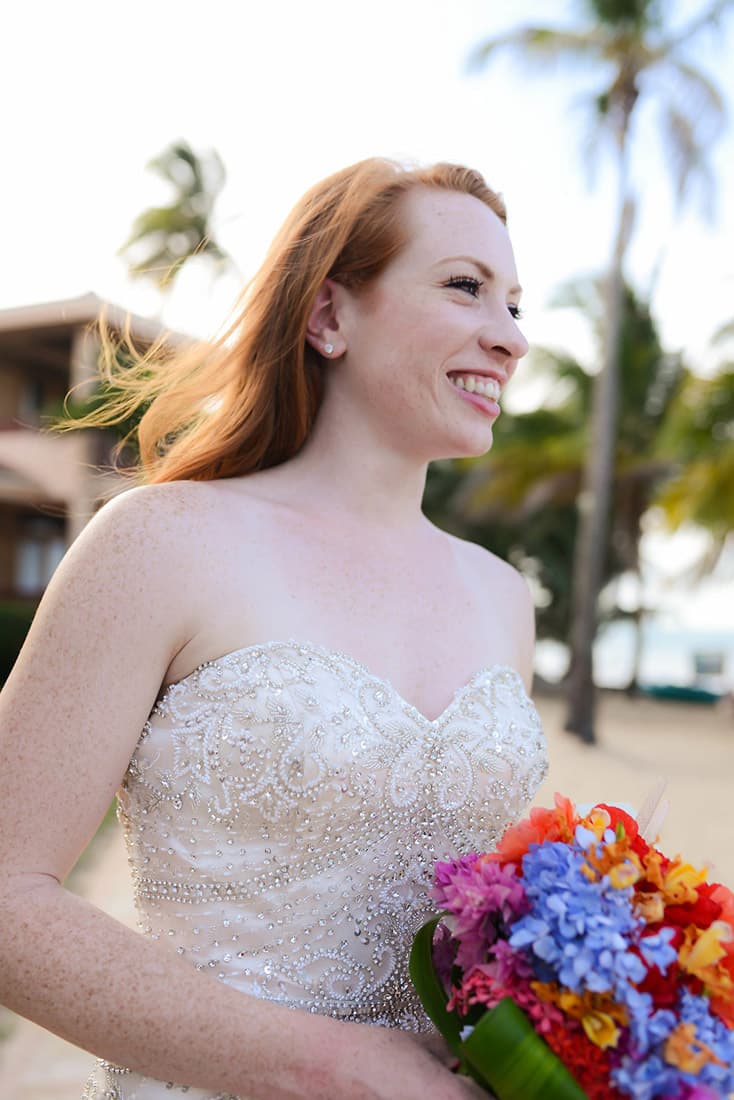 Beautiful red-headed bride in a strapless beaded gown stands on the beach with a colorful bouquet.