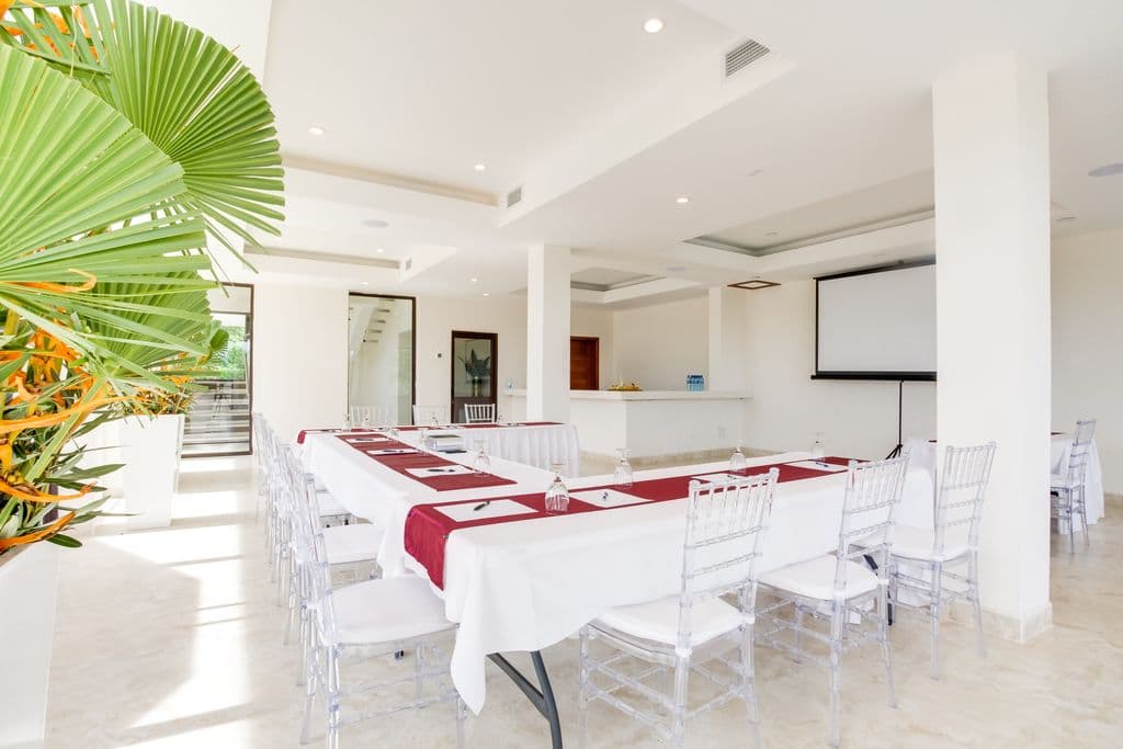 A spacious and modern meeting room with a U-shaped table arrangement. The room has a white tiled floor, white walls, and a high ceiling with exposed beams. There is a large palm tree in the corner, and a projection screen and projector at the front of the room. The table is covered in white tablecloths and has red runners. Clear plastic chairs are arranged around the table. There is a bar area with counter stools in the background.