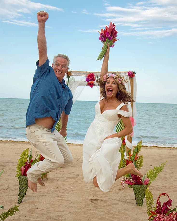 A mature bride and groom jump for joy in front of a wedding arch on the beach.