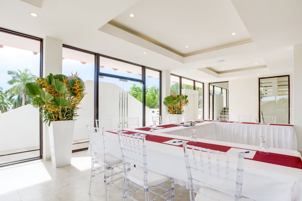 A modern meeting room with a U-shaped table arrangement. The room has a white tiled floor, white walls, and a high ceiling with exposed beams. A large projection screen and projector are at the front of the room. The table is covered in white tablecloths with red runners. Clear plastic chairs are arranged around the table. There is a bar area with counter stools in the background.