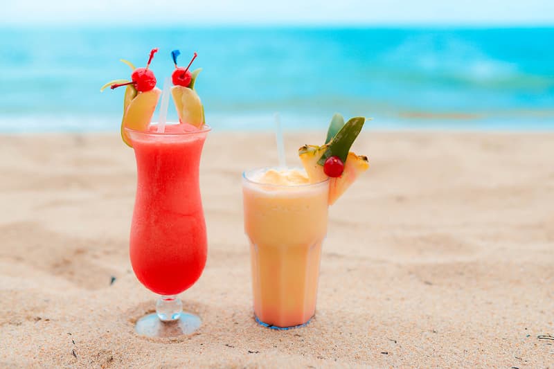 Two colorful tropical cocktails with garnishes sit on a sandy beach, with the ocean in the background. Two colorful tropical cocktails with garnishes sit on a sandy beach, with the ocean in the background.