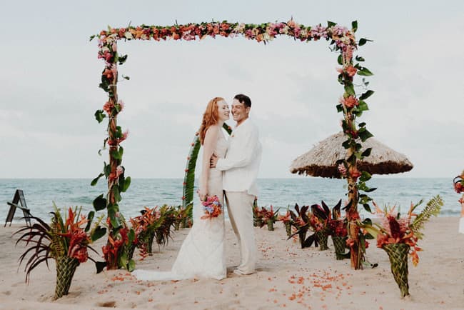 A smiling couple stands under a floral arch on the beach, surrounded by tropical plants and a thatched umbrella in the background.