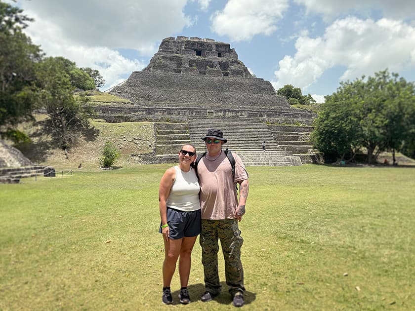 A couple stands in front of a large ancient pyramid surrounded by greenery under a partly cloudy sky. A couple stands in front of a large ancient pyramid surrounded by greenery under a partly cloudy sky.