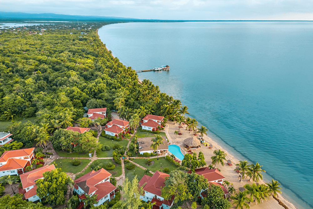 An aerial view of Belizean Dreams Resort, showing a cluster of white buildings with red roofs nestled along a sandy beach. The resort is surrounded by lush green vegetation and overlooks a calm blue ocean. There is a pier extending into the water and a swimming pool visible within the resort grounds.