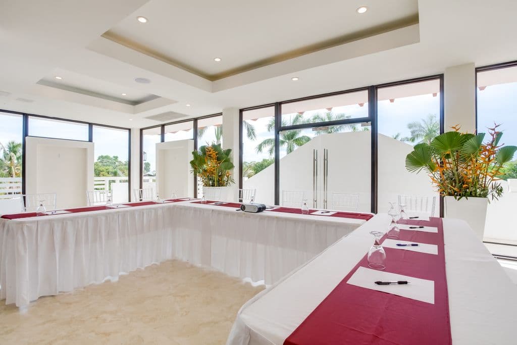 A modern meeting room with a U-shaped table arrangement. The room has a white tiled floor, white walls, and a high ceiling with exposed beams. Large windows offer views of a green outdoor space. The table is covered in white tablecloths with red runners. Clear plastic chairs are arranged around the table. There are floral arrangements in white vases on either side of the room.