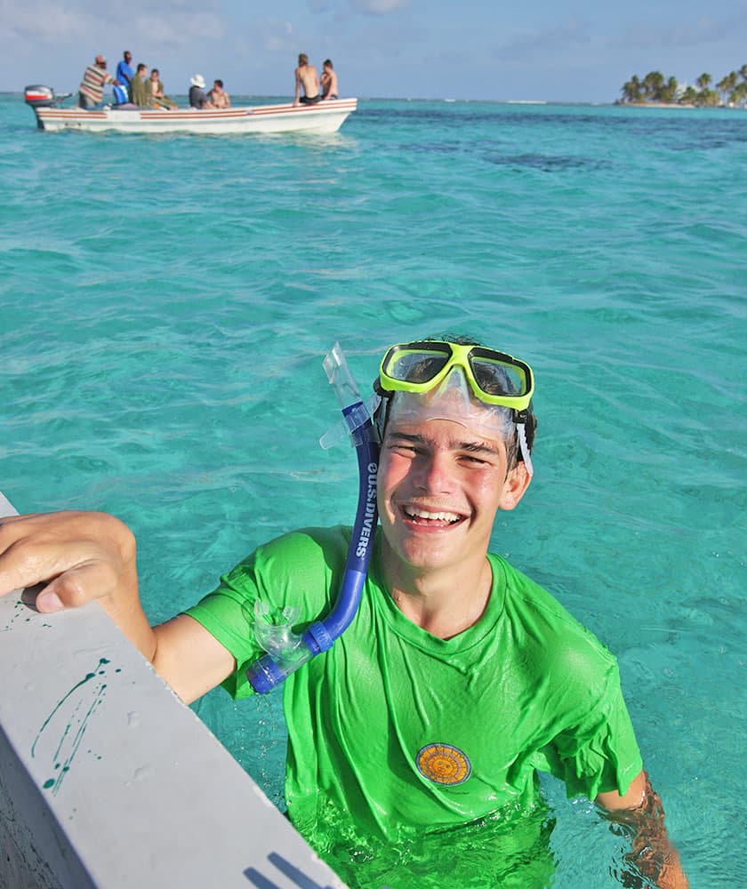 A joyful boy in a green shirt and snorkeling gear smiles while emerging from turquoise water, with a boat and people in the background.