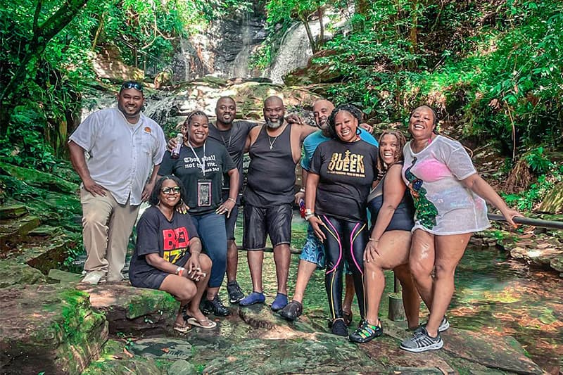 A group of nine people stands together by a waterfall in a lush, green setting. A group of nine people stands together by a waterfall in a lush, green setting.