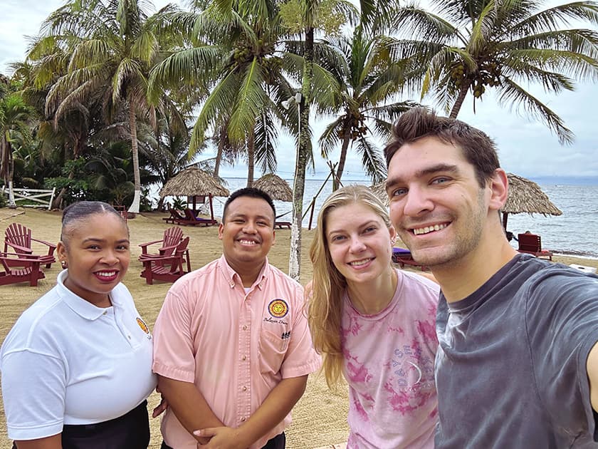 A group of four people smiles for a selfie on a beach with palm trees and traditional huts in the background.
