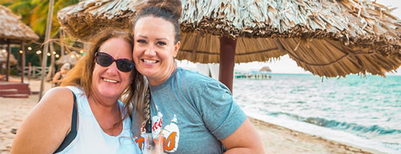 Two smiling women pose together on a beach under a straw umbrella. Two smiling women pose together on a beach under a straw umbrella.