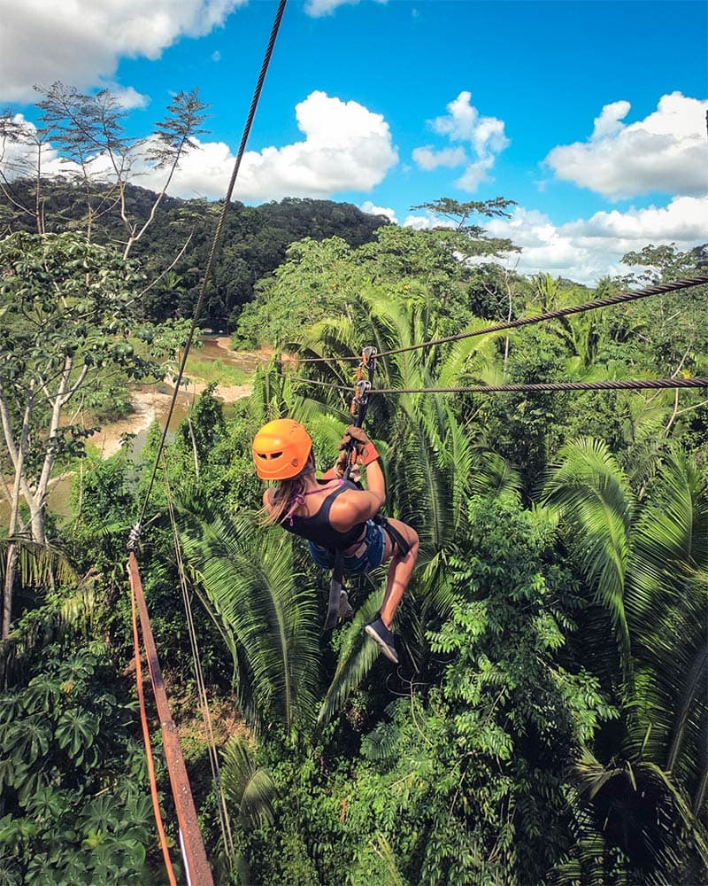A person in an orange helmet zip-lines through a lush green forest under a blue sky.