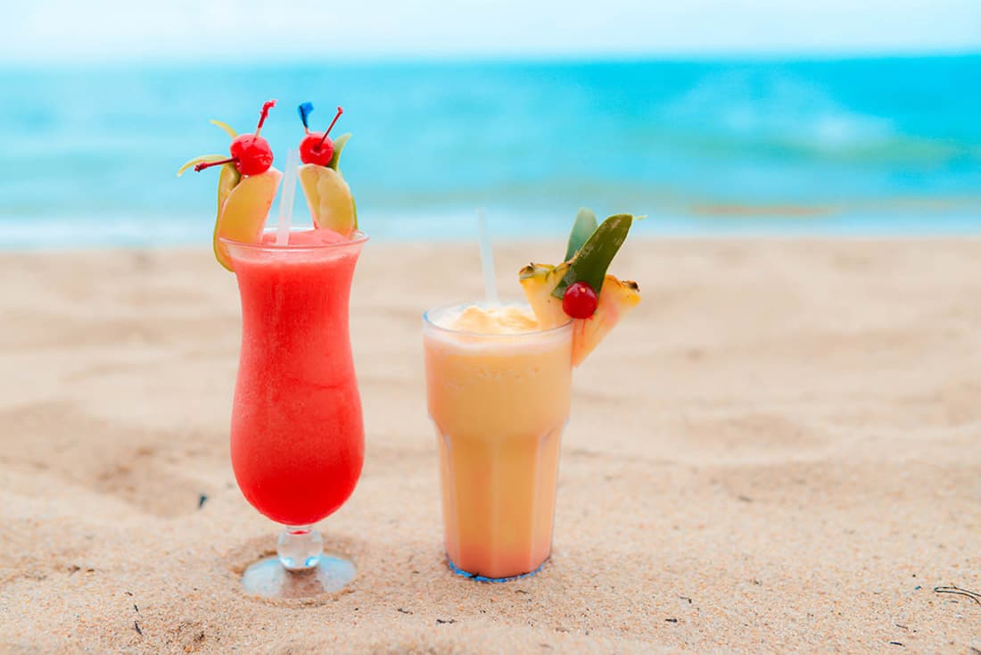 Two colorful tropical drinks on a sandy beach. The left drink is a bright red color, possibly a strawberry daiquiri, with a pineapple wedge and cherries on the rim. The right drink is a lighter orange color, possibly a piña colada, with a pineapple wedge and a cherry on the rim. Both drinks are served in tall glasses with straws. The ocean is visible in the background.