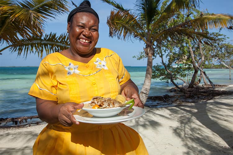 A smiling woman in a yellow dress holds a plate of food on a beach with palm trees and the ocean in the background.