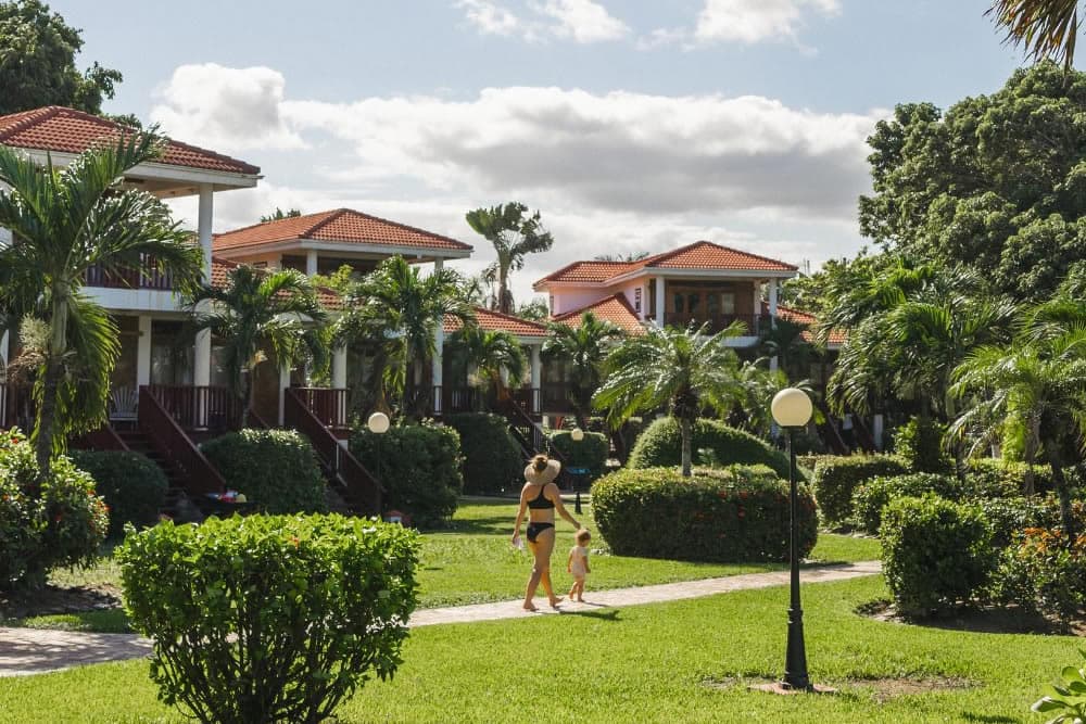 A woman in a black swimsuit and sunhat walks with a toddler down a path, surrounded by greenery and white villas with red tile roofs.