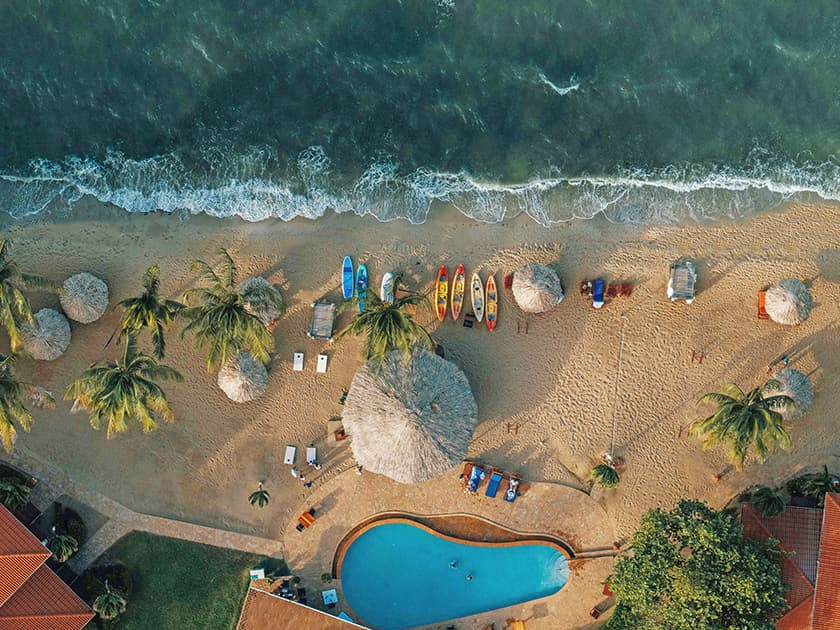 Aerial view of a sandy beach with palm trees, umbrellas, a swimming pool, and colorful kayaks. Aerial view of a sandy beach with palm trees, umbrellas, a swimming pool, and colorful kayaks.