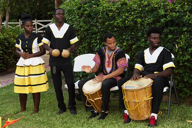 A group of four musicians in traditional attire perform with drums and maracas in a lush outdoor setting.