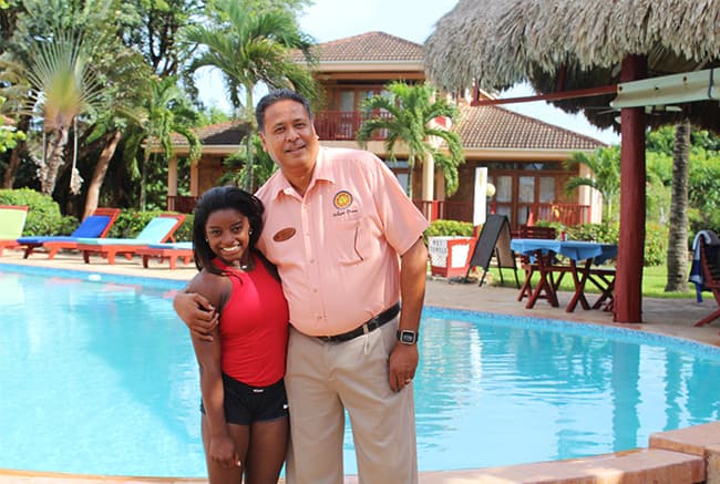 A smiling woman in a red top and a man in a pink shirt pose together by a pool surrounded by lush greenery and colorful lounge chairs.